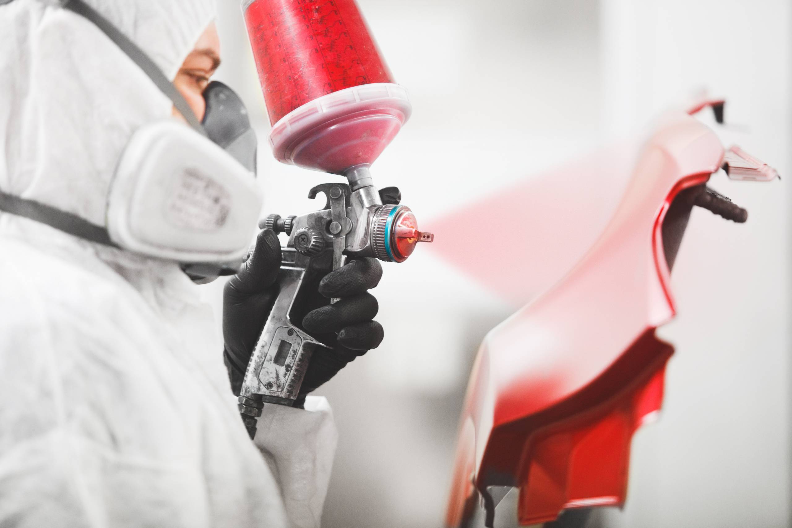 A person in a white protective suit and respirator uses a spray gun to paint a car bumper red for an auto body repair.