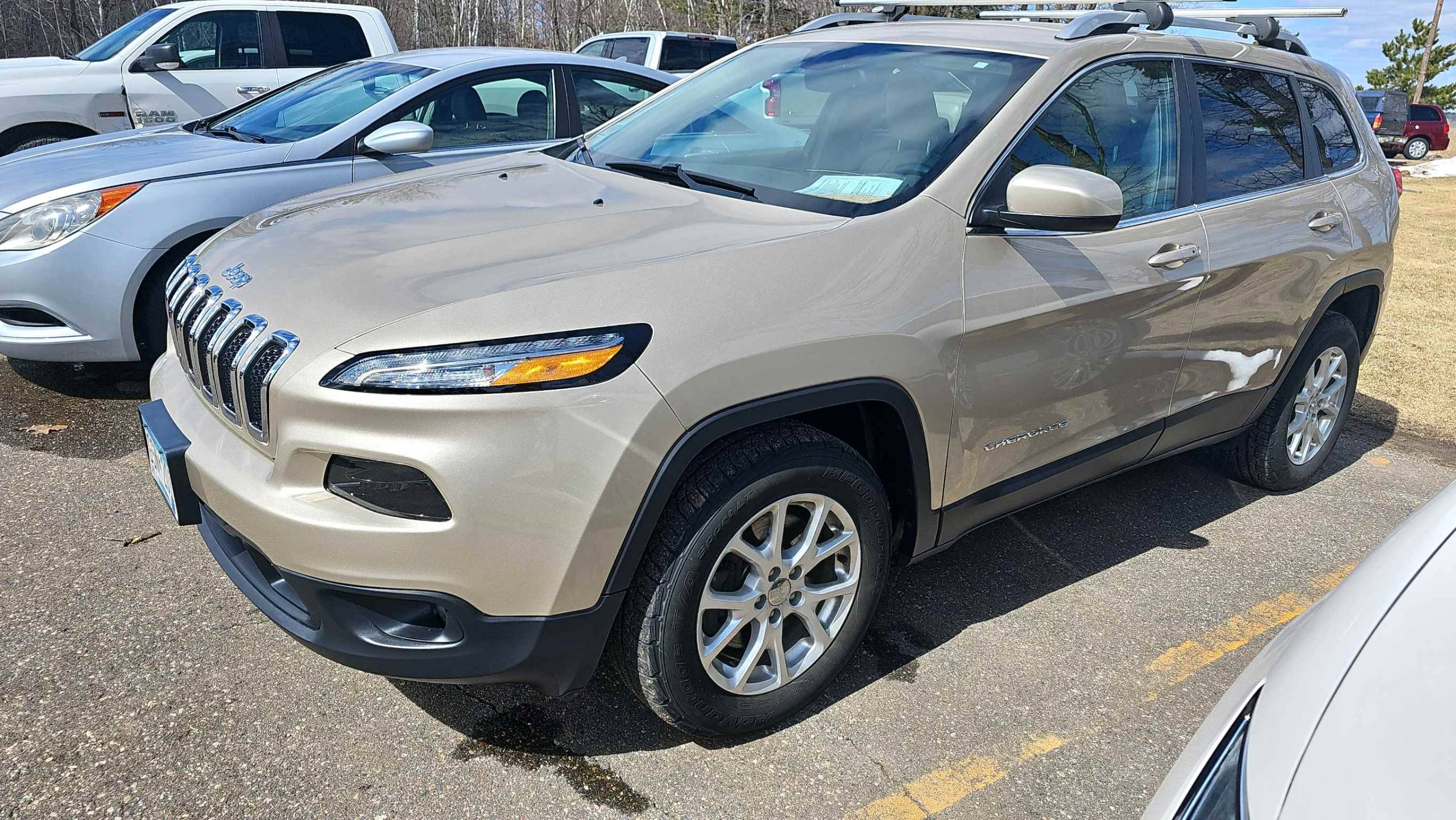 A tan Jeep Cherokee undergoes auto body work in a lot next to a silver car.