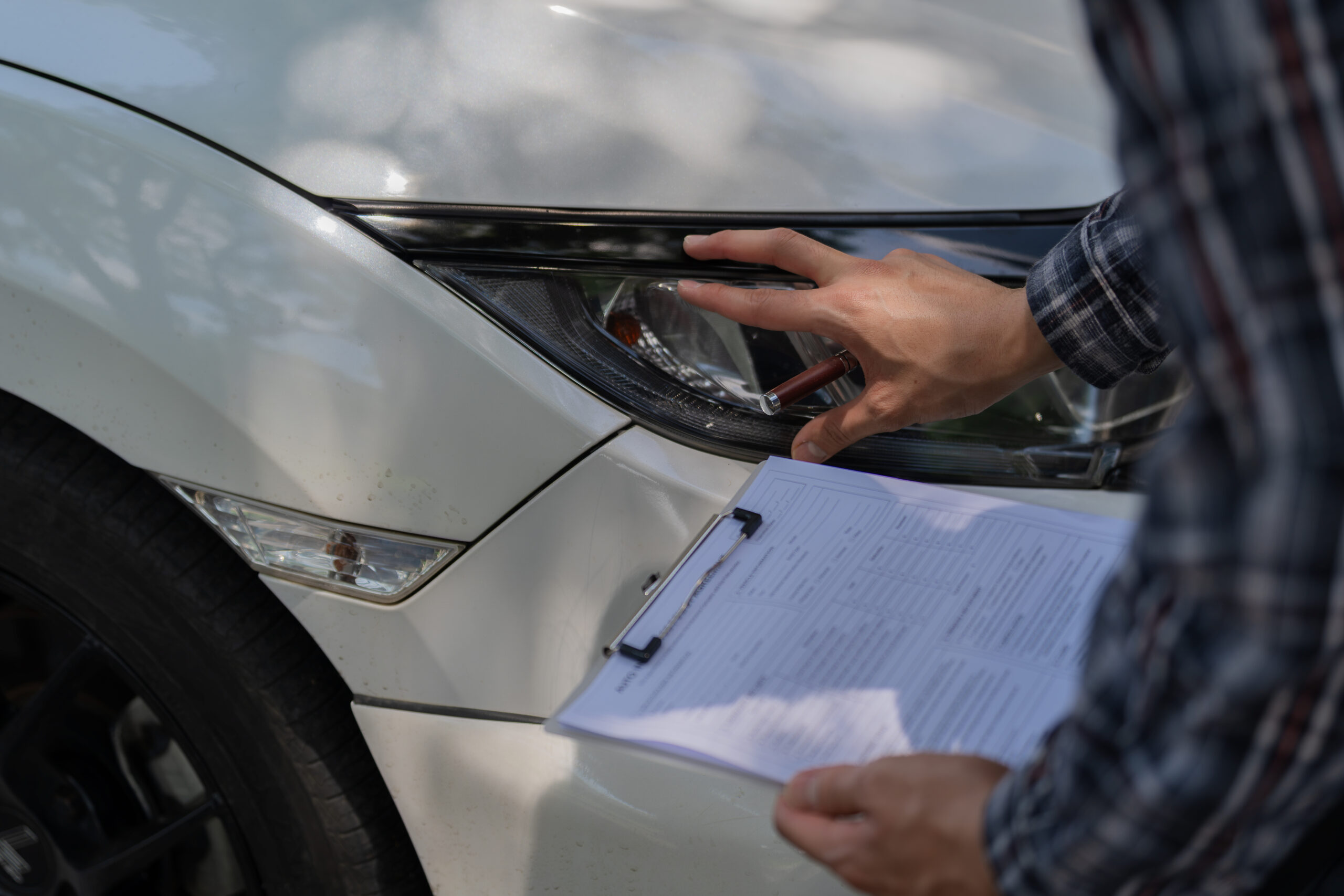 A person holding a clipboard points at the headlight of a white car during a collision repair assessment.